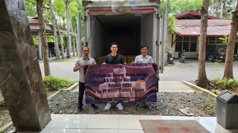 Tiga orang memegang spanduk pelepasan ekspor cocoa butter di depan truk boks yang terbuka, kardus barang terlihat di dalam truk.
